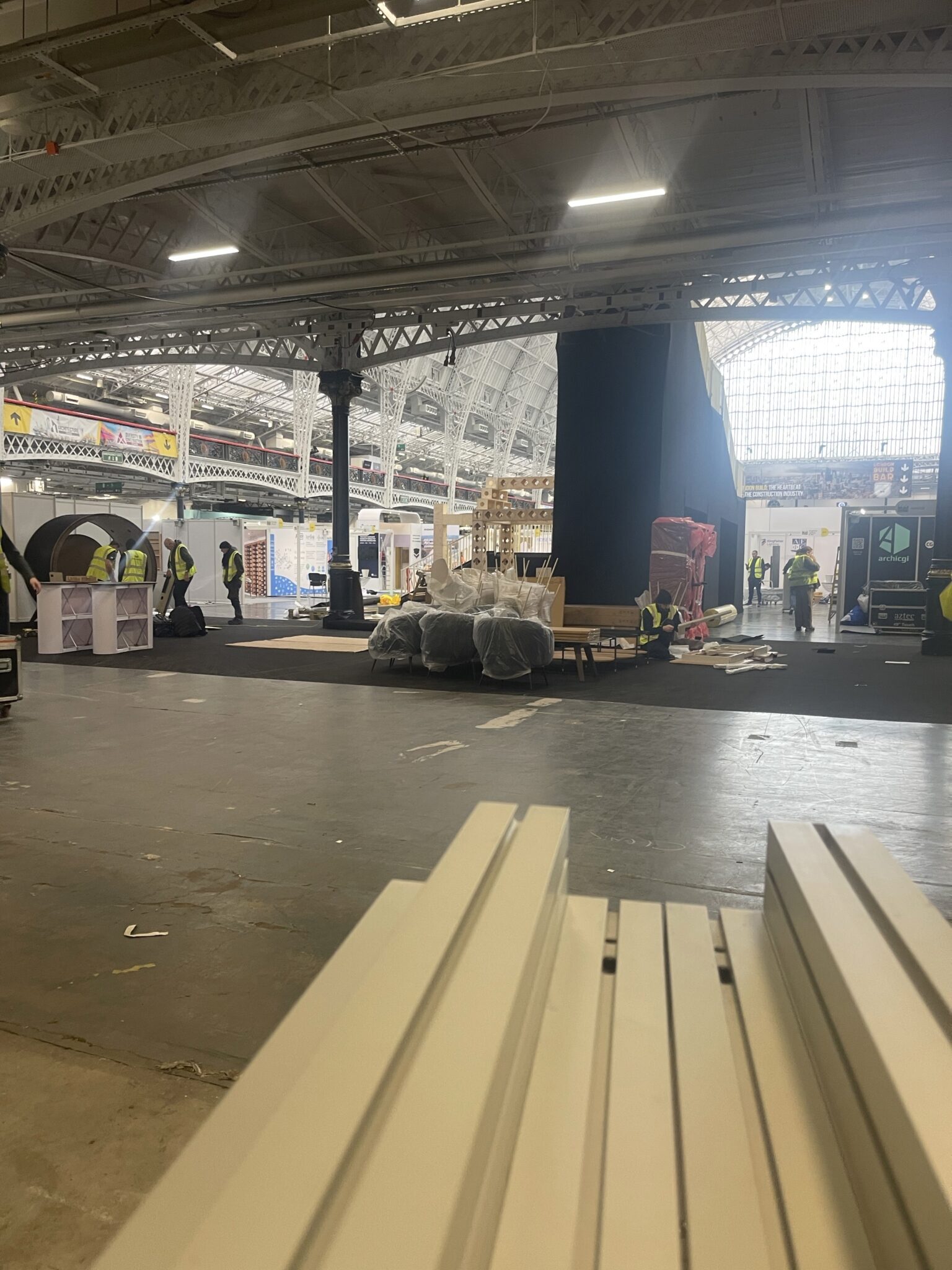 Wide view of London Olympia exhibition hall showing ESV Removals unloading stand materials for Sigicom, with pallets, wrapped displays and crew in high visibility vests preparing for set up.